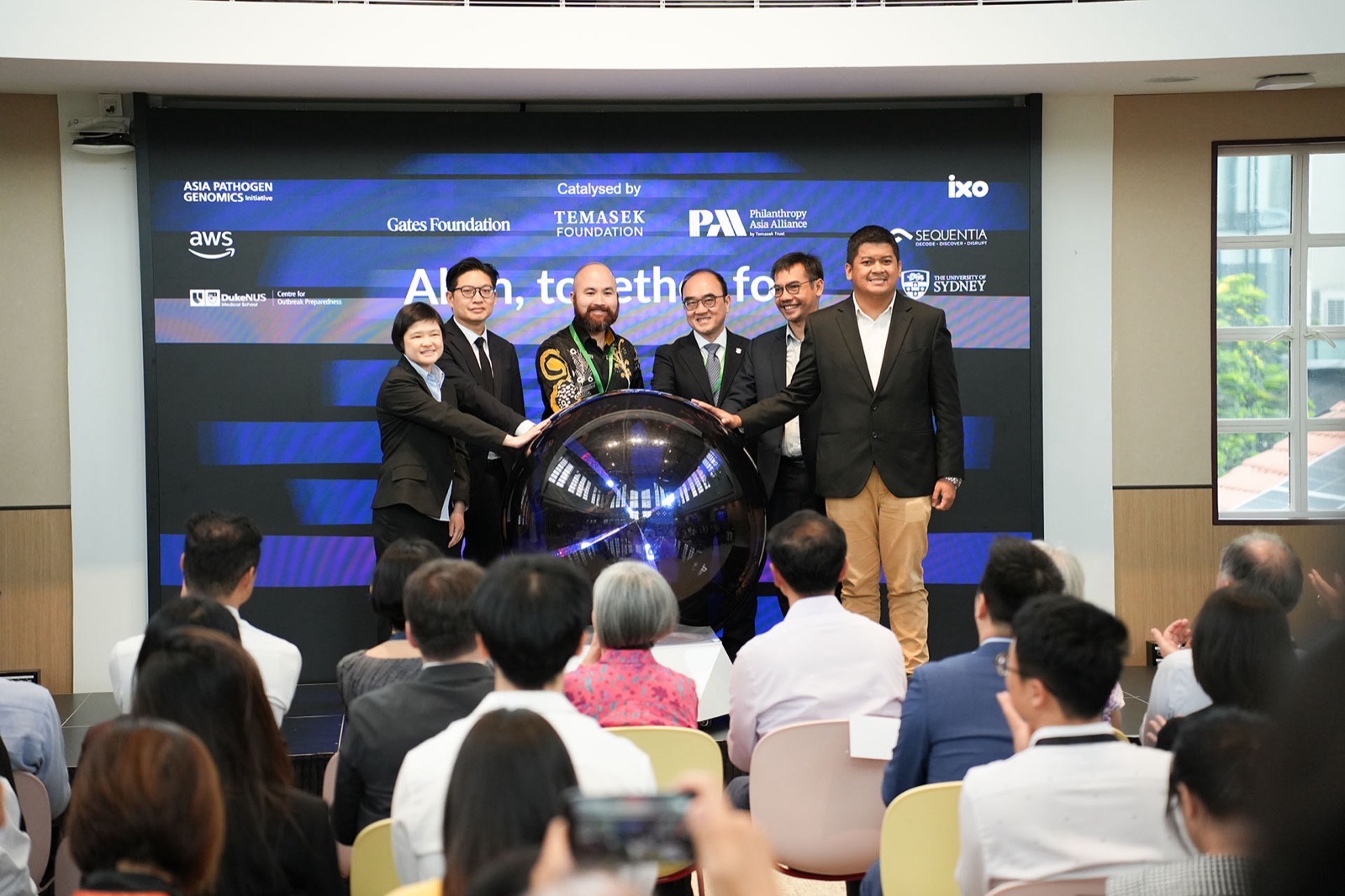 Six people standing side by side with their hands on a mirrored sphere on stage, in front of the PathGen preview backdrop, as the audience watches.