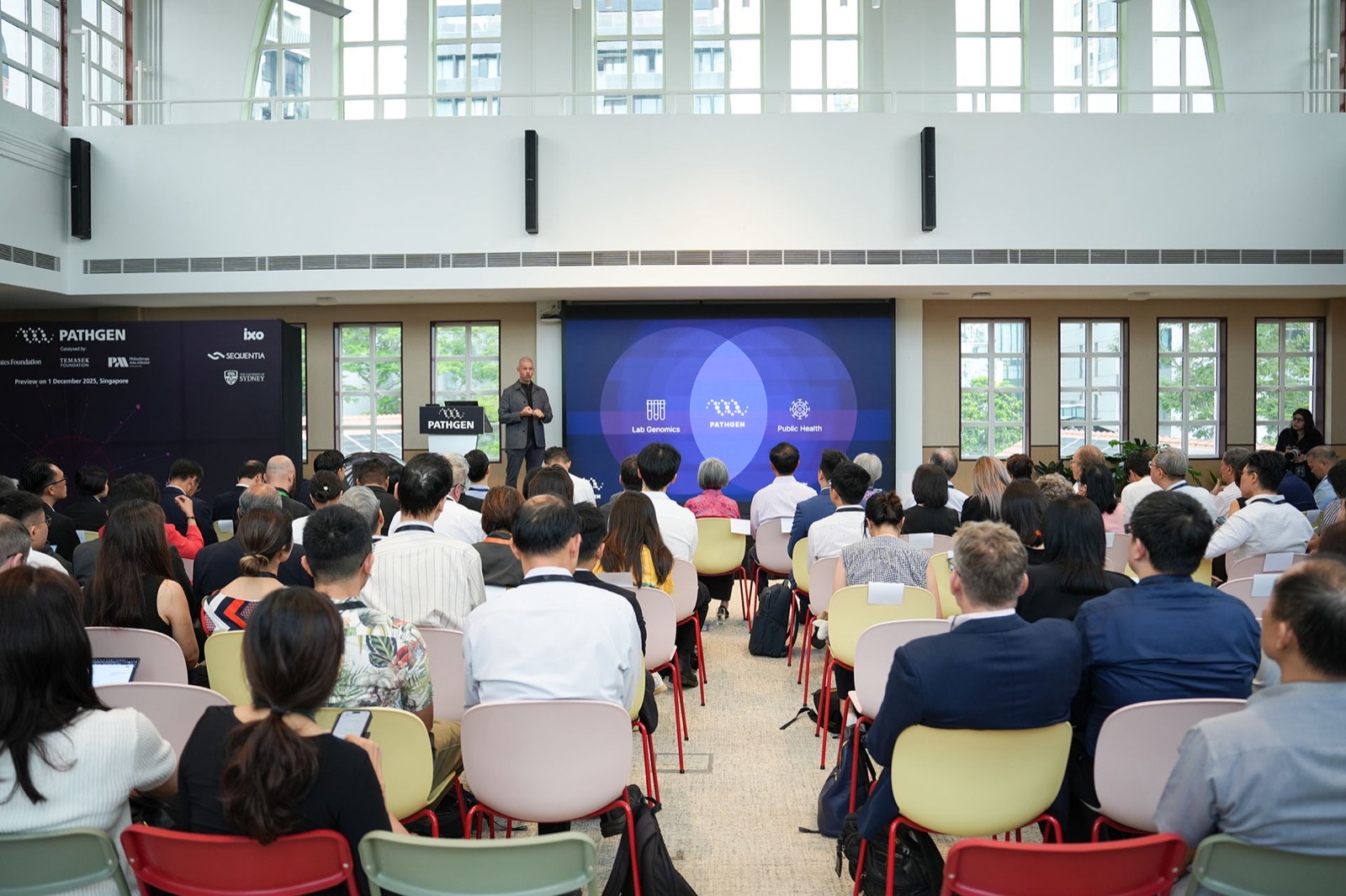 A seated audience filling a naturally lit hall, facing a speaker beside a screen showing three overlapping circles labelled lab genomics, PathGen and public health.