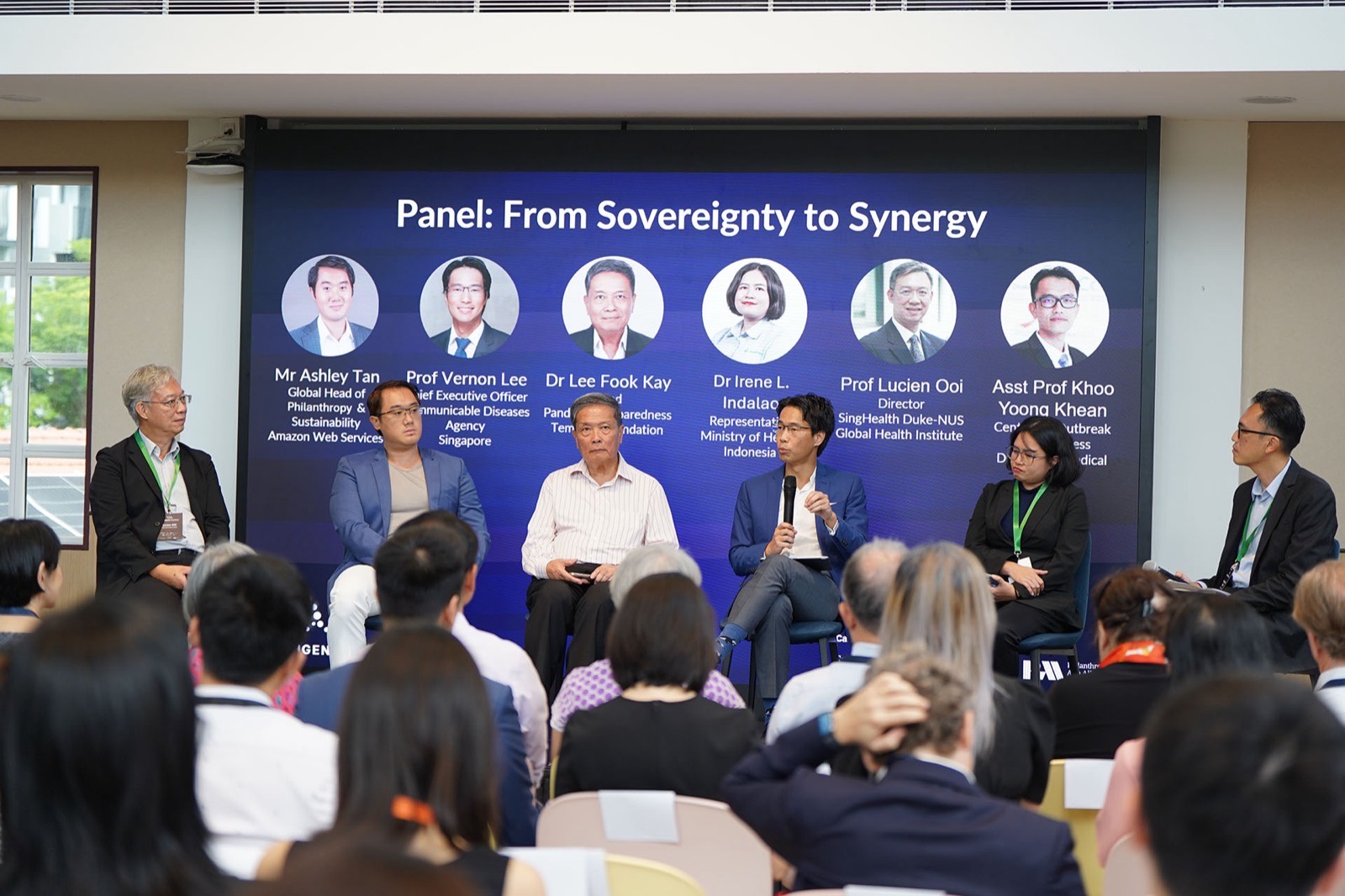 Seven people seated on a low stage for a panel discussion, in front of a screen headed “Panel: From Sovereignty to Synergy”.